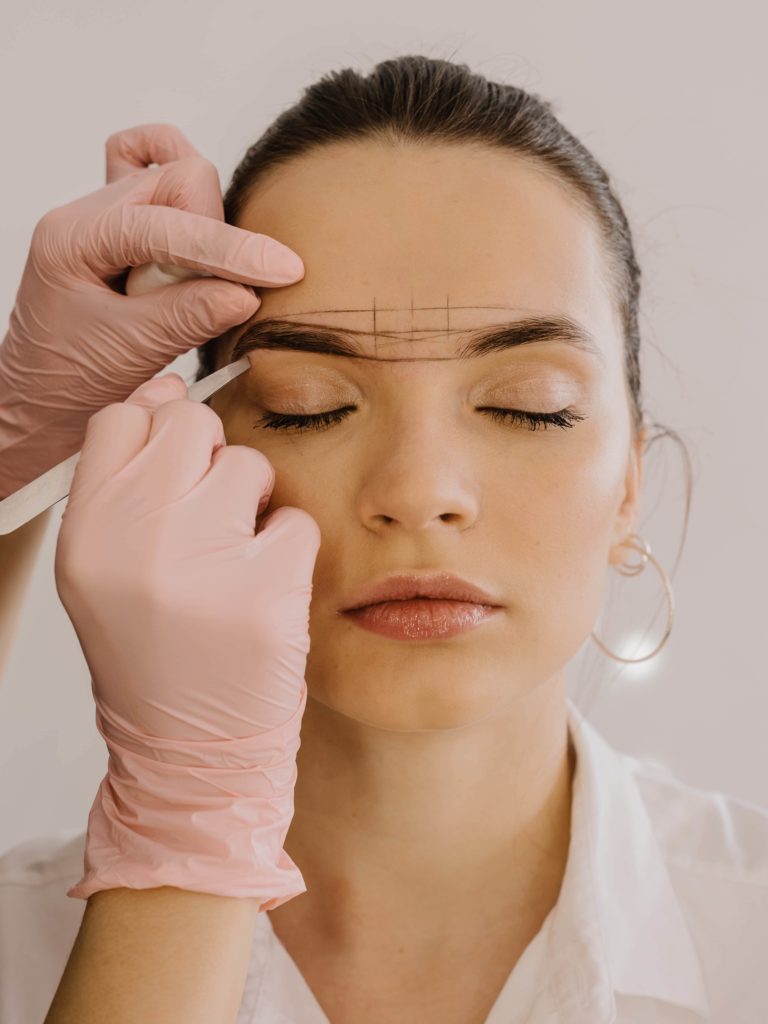 Woman receiving eyebrow mapping for microblading; technician in pink gloves marking brows with measuring lines