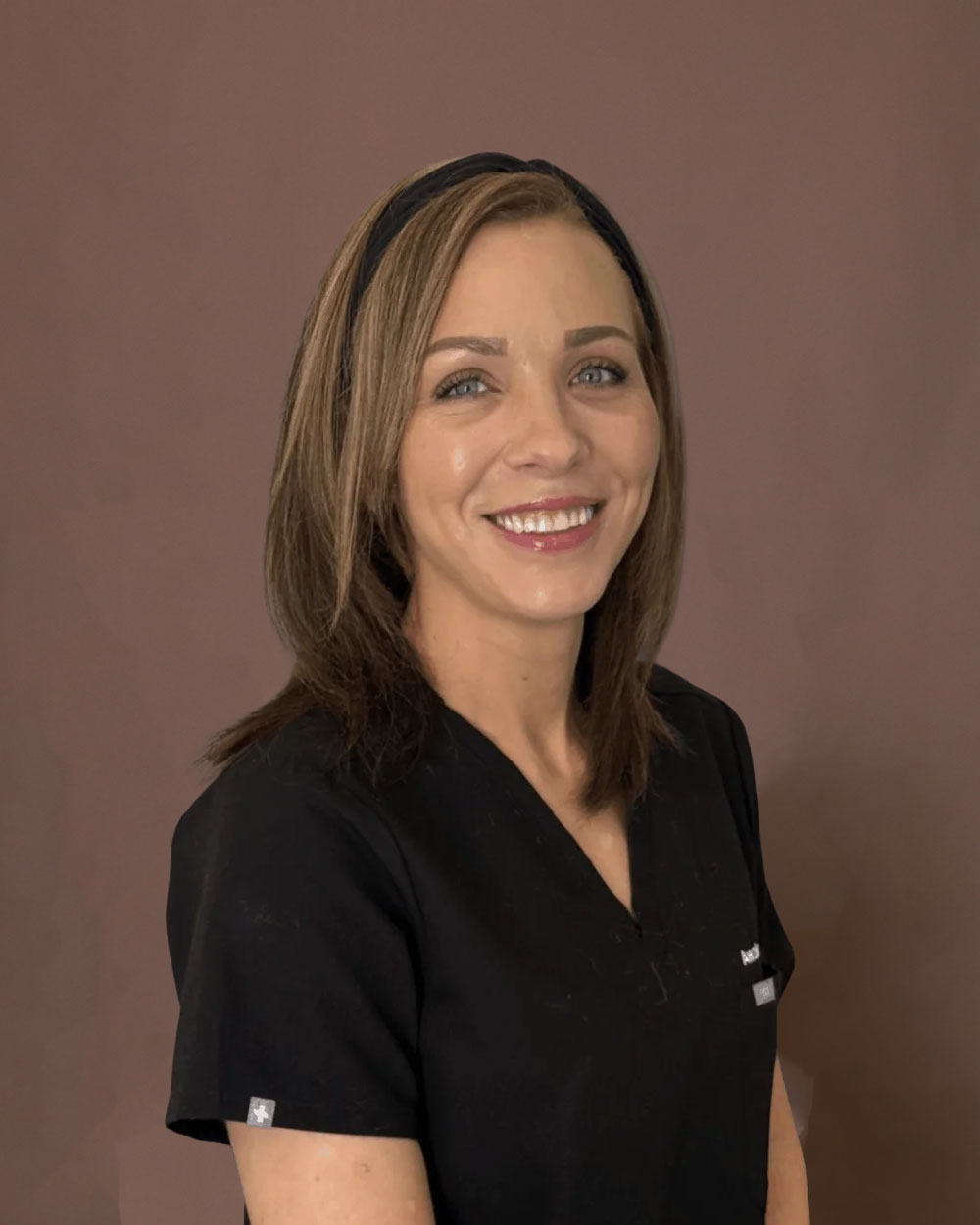 A smiling woman in black medical scrubs with a headband against a brown background.