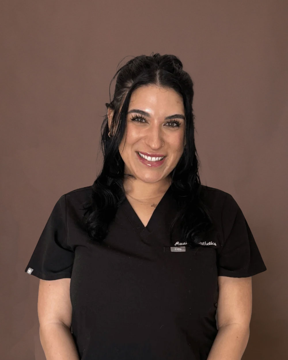 A smiling woman in black medical scrubs in front of a brown backdrop, professional headshot