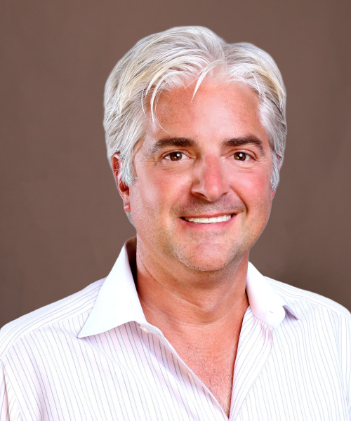 Smiling middle-aged man with silver hair in white collared shirt, professional headshot on plain brown background.