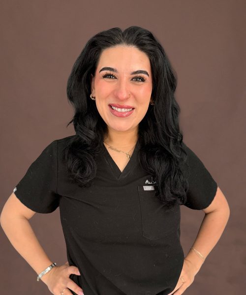 Smiling woman in black medical scrubs with hands on hips, long dark hair against a brown background
