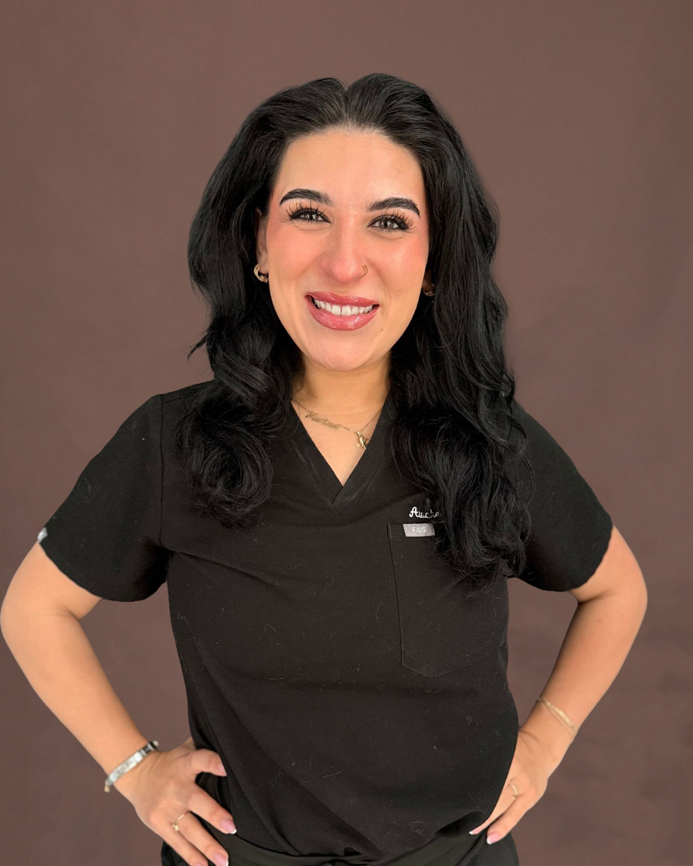 Smiling woman in black medical scrubs with hands on hips, long dark hair against a brown background
