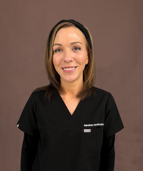 Smiling woman in black medical scrubs with 'Awaken Aesthetics' logo on chest against brown backdrop.
