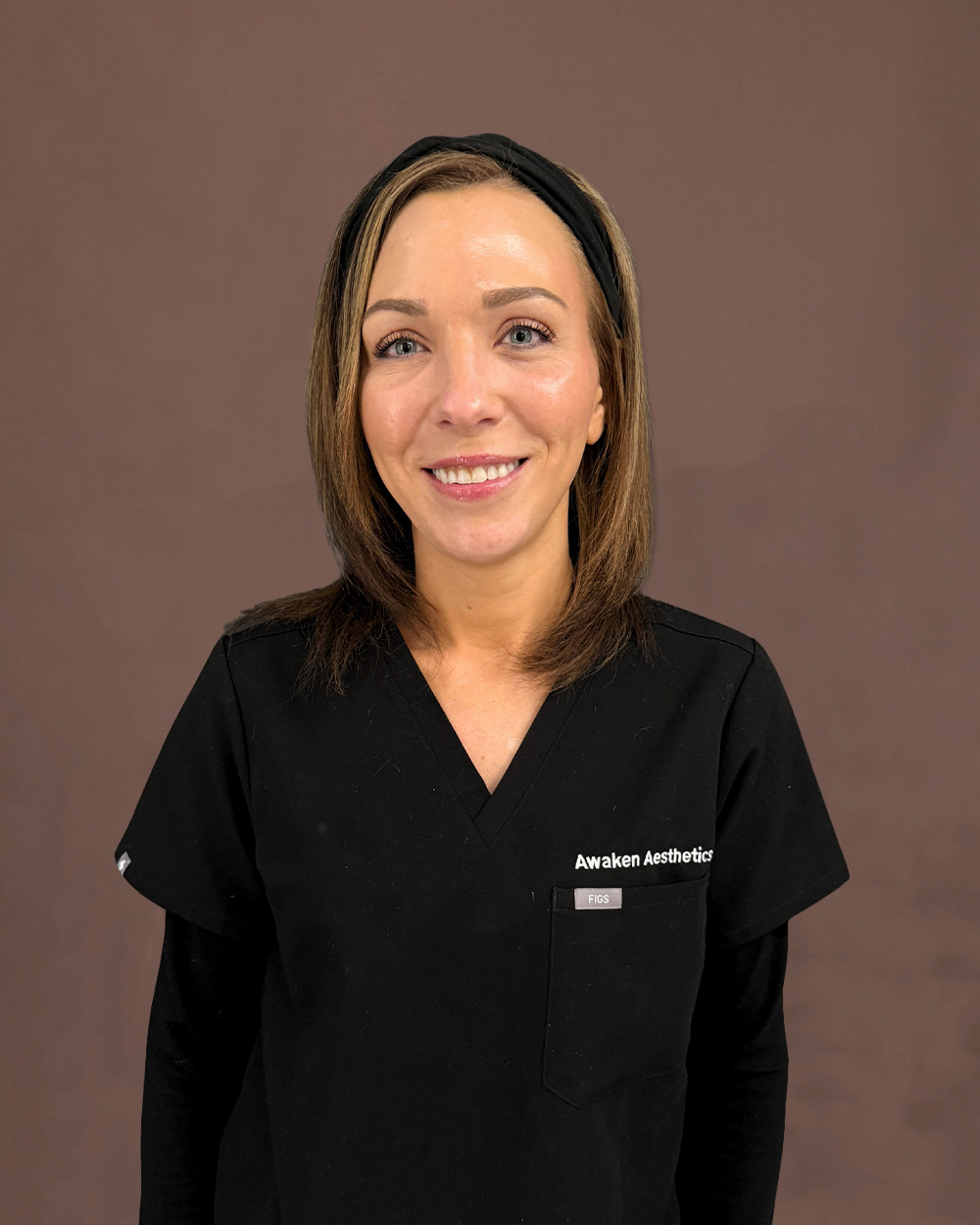 Smiling woman in black medical scrubs with 'Awaken Aesthetics' logo on chest against brown backdrop.