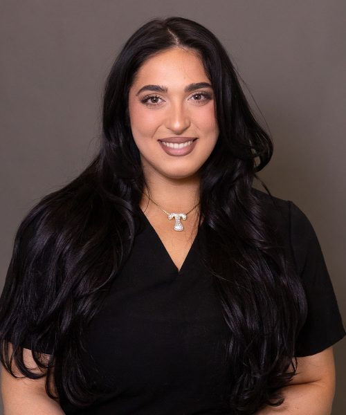 A woman with long dark hair in a black top smiling at the camera, wearing a gold T-shaped pendant necklace