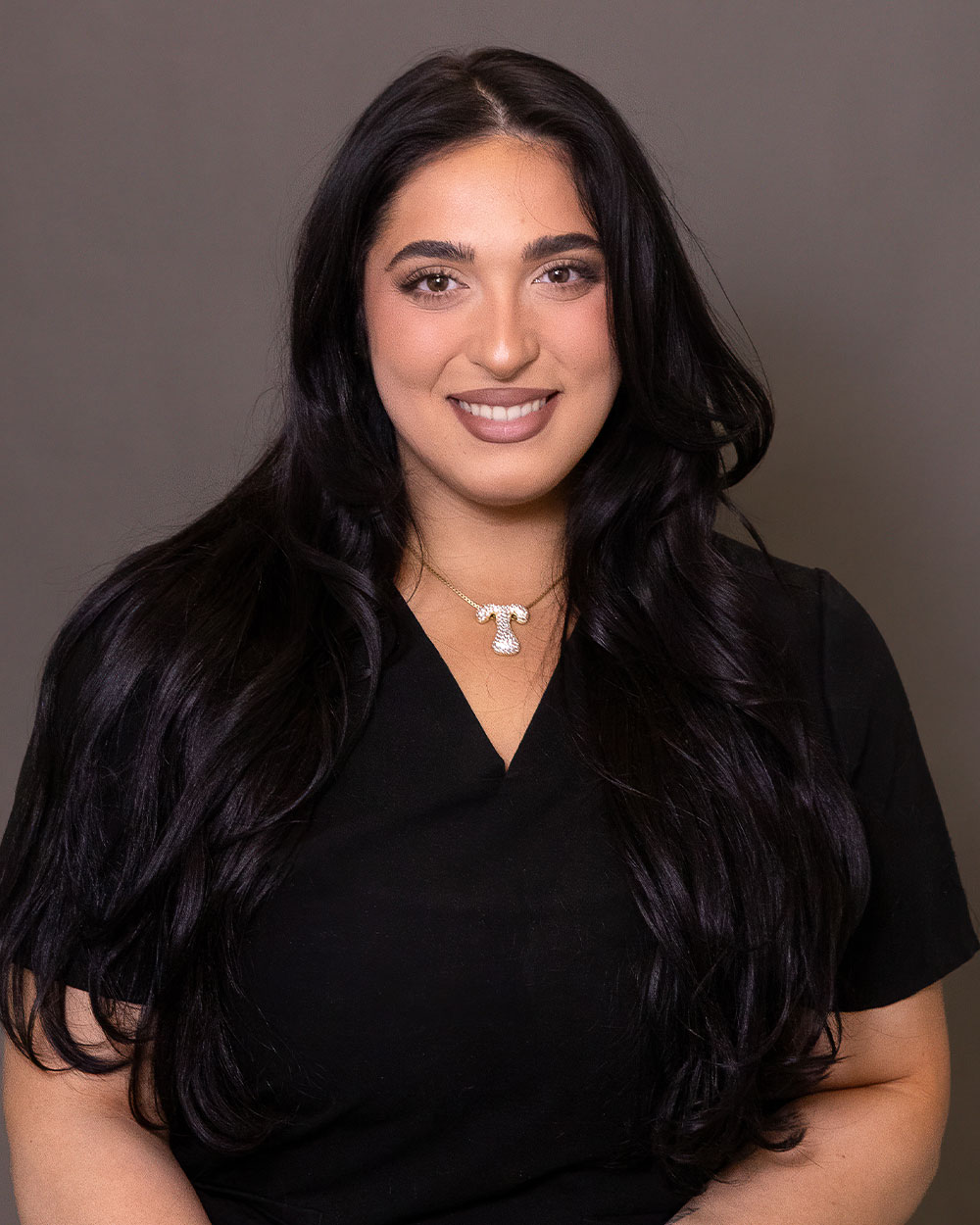 A woman with long dark hair in a black top smiling at the camera, wearing a gold T-shaped pendant necklace