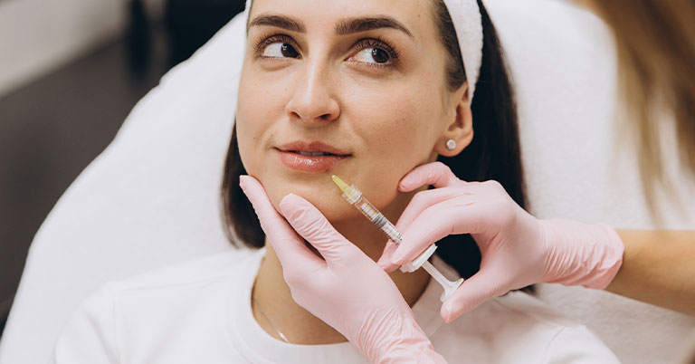 A woman receiving a cosmetic lip filler injection from a clinician wearing pink gloves in a medical spa.