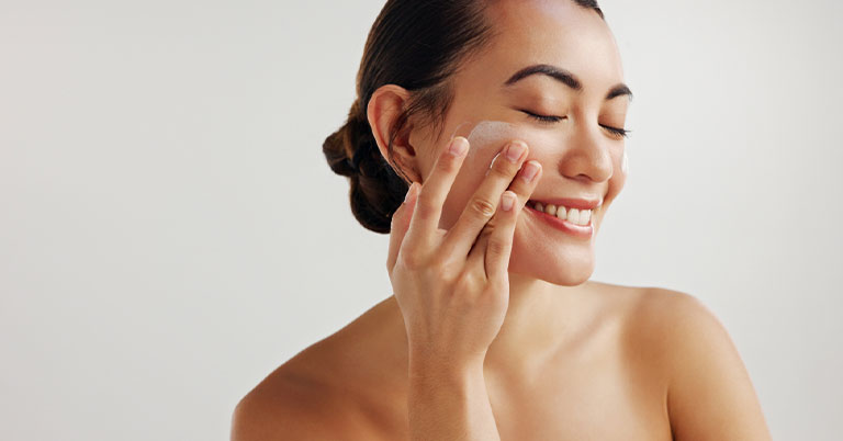 Woman applying facial cream with a smile, part of a skincare routine for glowing, hydrated skin.