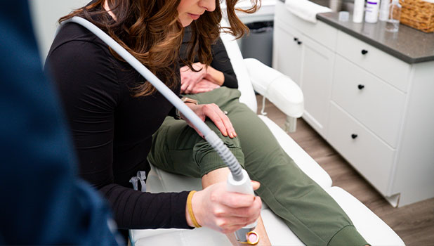 A woman performing laser hair removal on a patient's lower leg in a modern medical spa treatment room.