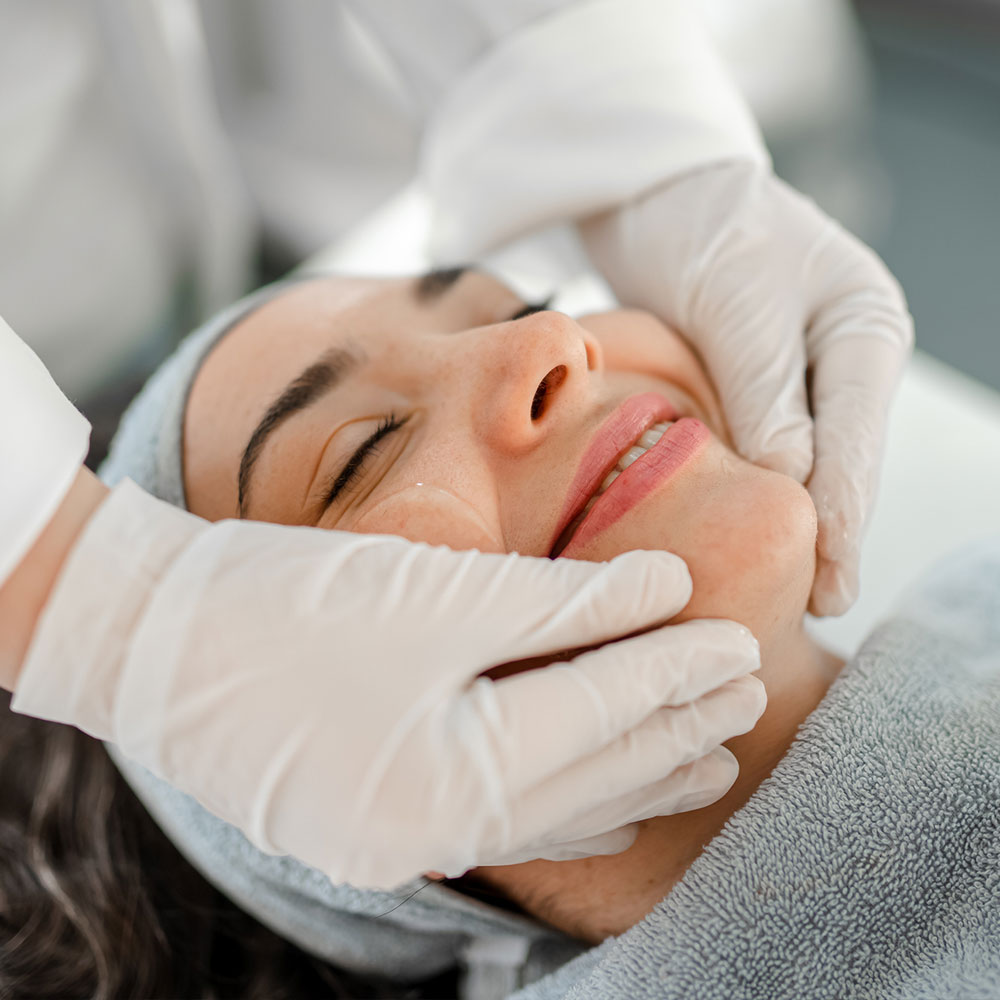 Close-up of a smiling woman receiving a relaxing facial treatment from a gloved esthetician
