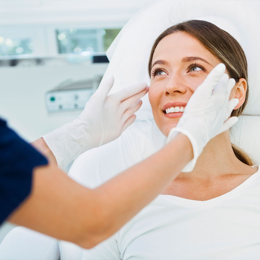 A woman smiling as a clinician wearing gloves examines her face in a cosmetic clinic.