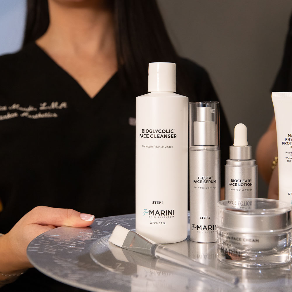 A woman holding a tray of Jan Marini skincare products, featuring Bioglycolic Face Cleanser, serum and face lotion.