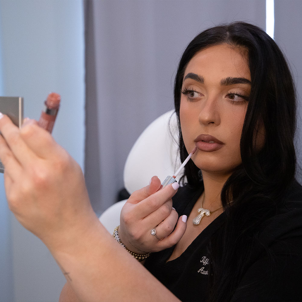 A woman applying nude lip gloss while looking into a handheld mirror, close-up makeup application