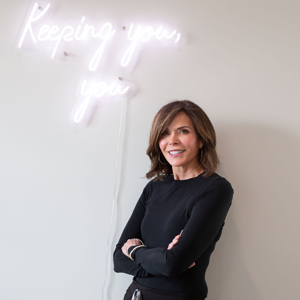 A smiling woman with arms crossed standing by neon sign reading Keeping you, you on a pale wall