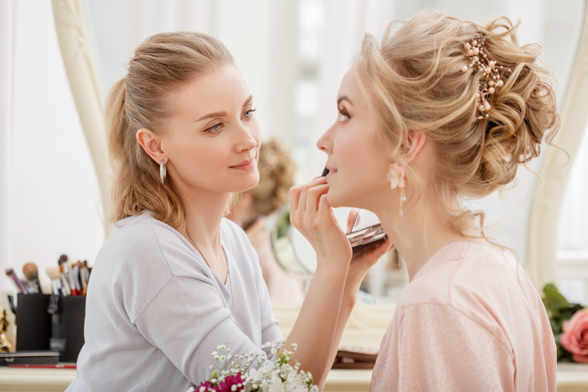 Makeup artist applying lipstick to a woman with a floral hairpiece and elegant updo, holding an eyeshadow palette.