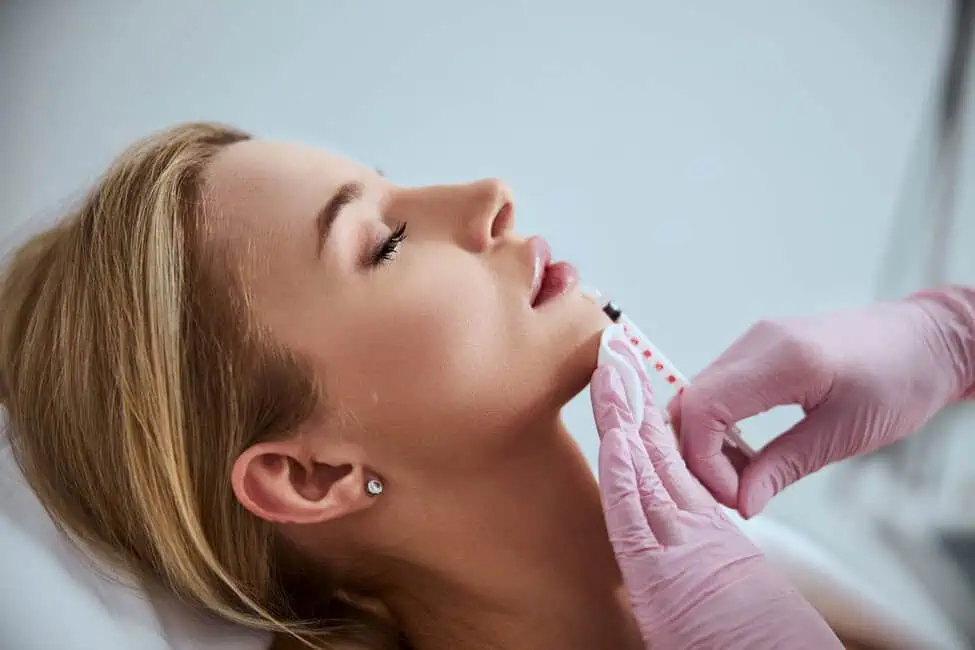 A woman receiving chin dermal filler injection from clinician wearing pink gloves