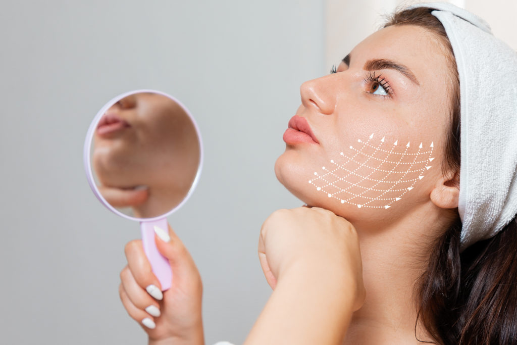 Woman holding mirror, examining chin while a dotted mesh with arrows on her cheek shows facial lifting and contouring.