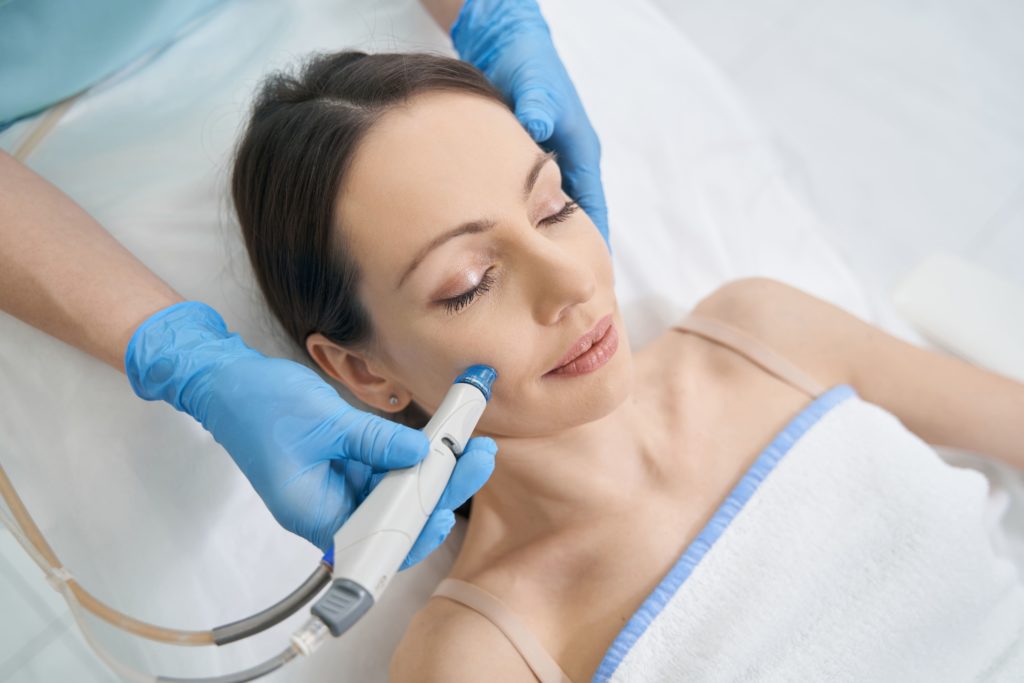 A woman receiving a hydrafacial skin treatment with a handheld device from a gloved esthetician while reclining.