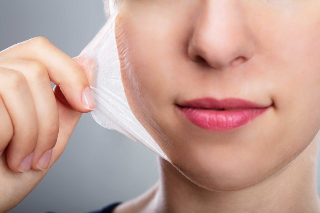 Close-up of a woman peeling a translucent facial peel-off mask from her cheek, showing smooth skin and pink lips.