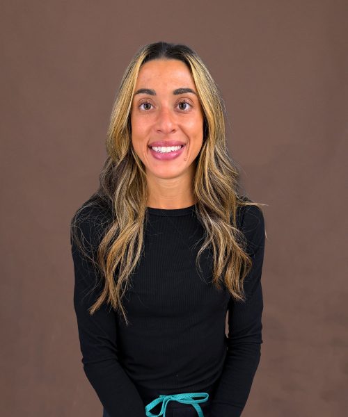 Smiling woman with long highlighted hair wearing a black top and teal drawstring against a brown backdrop
