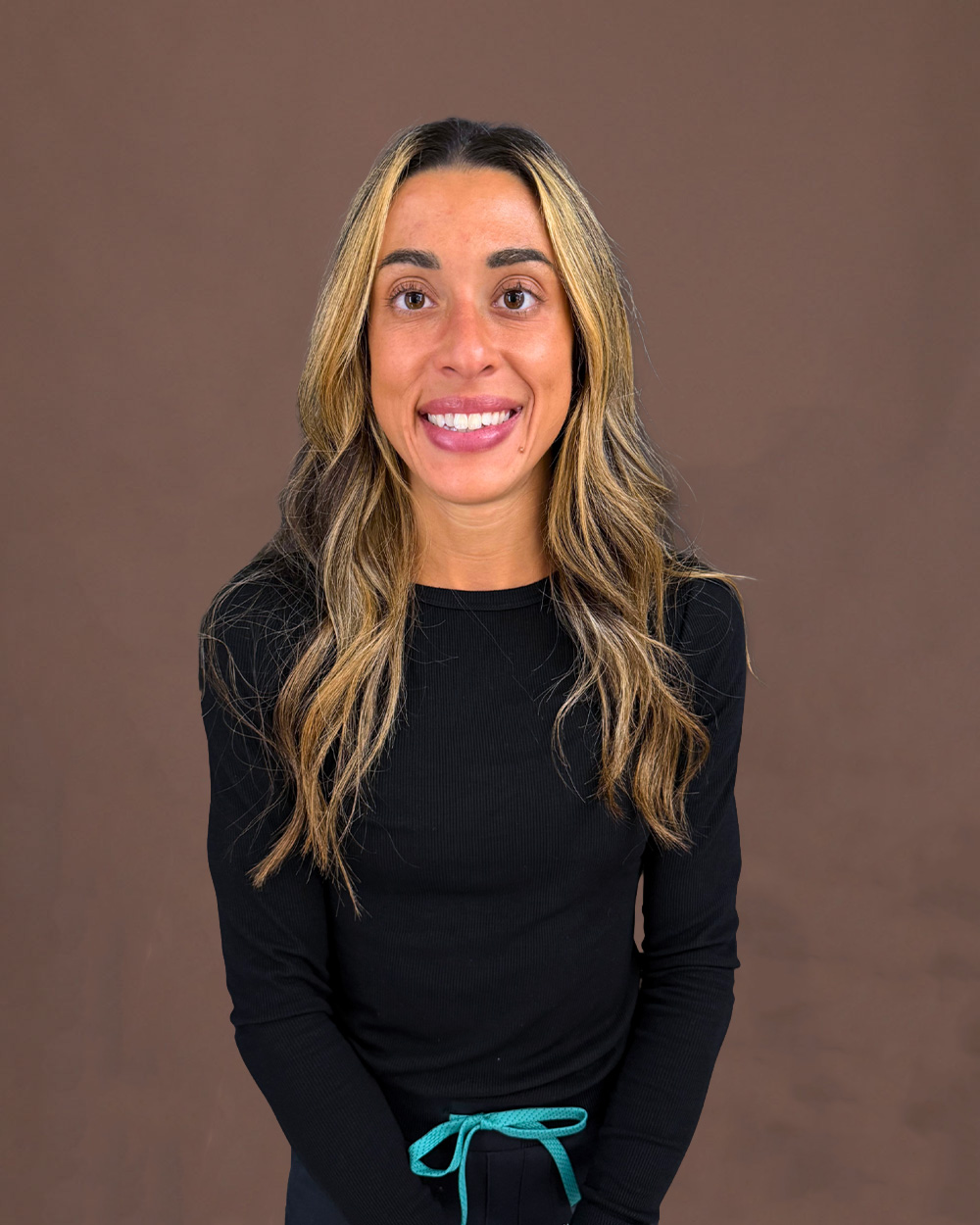 Smiling woman with long highlighted hair wearing a black top and teal drawstring against a brown backdrop