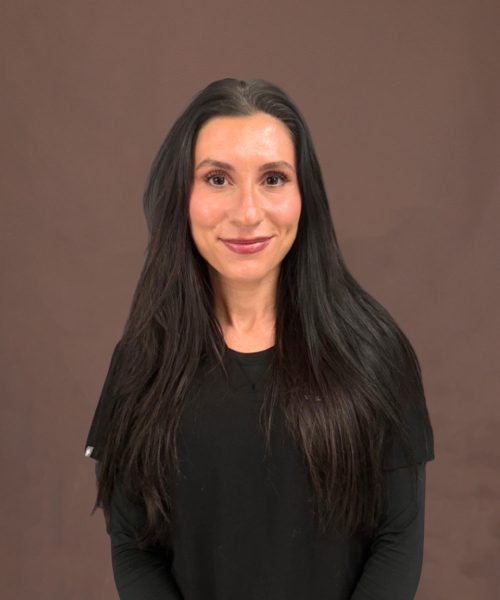 Portrait of a woman with long dark hair wearing a black top, smiling against a neutral brown background.