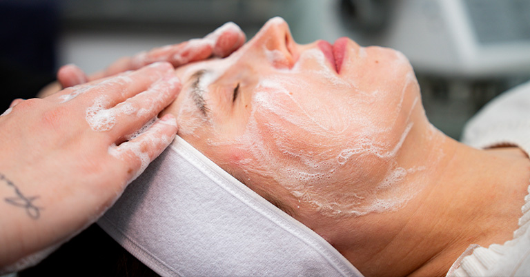 Woman lying with cleansing foam on face while esthetician massages her forehead during a facial treatment