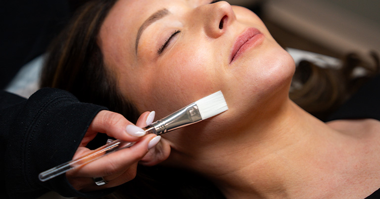 Close-up of woman receiving facial treatment as esthetician applies mask with brush to her jaw and chin.