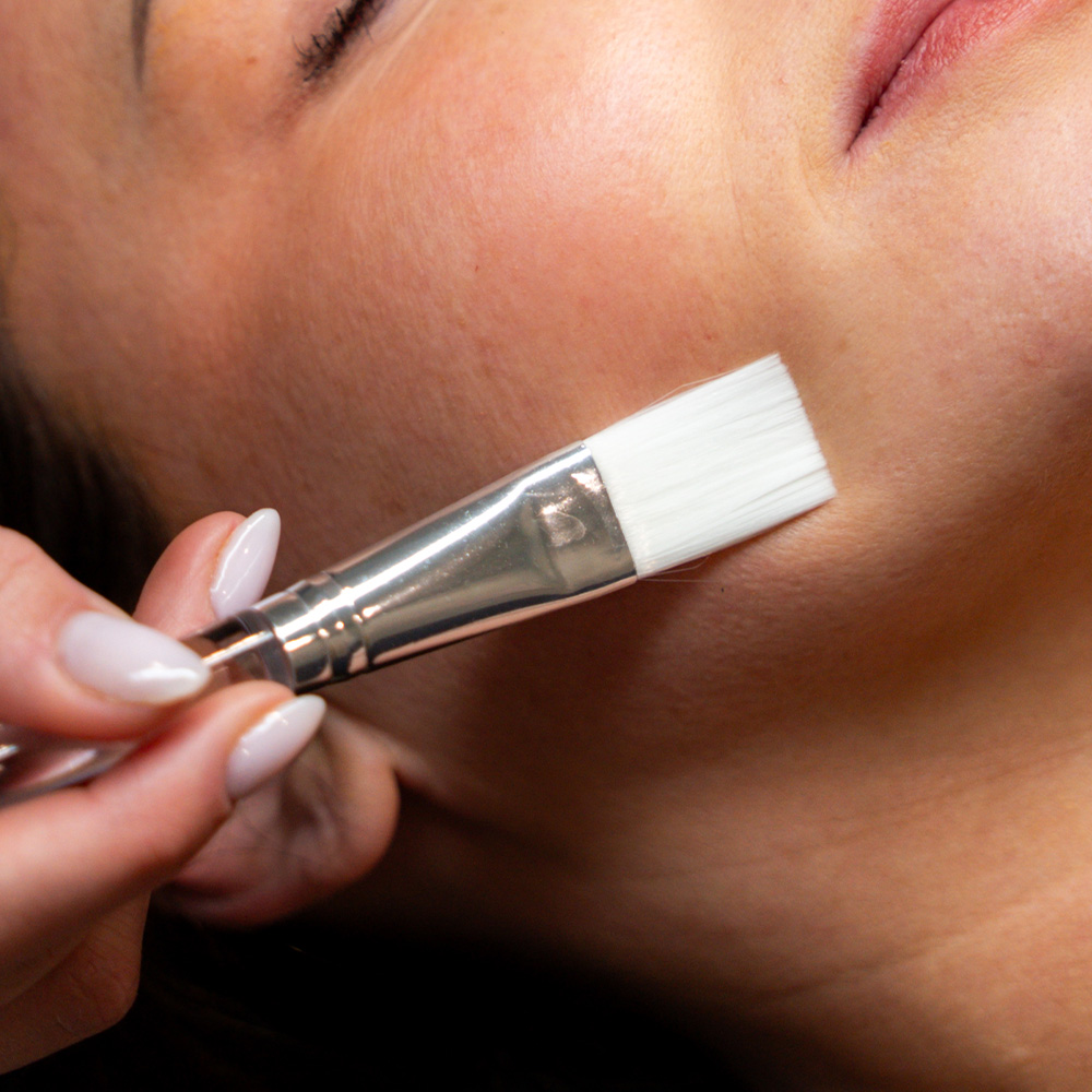 A woman receiving a white facial mask applied to her cheek with a flat brush