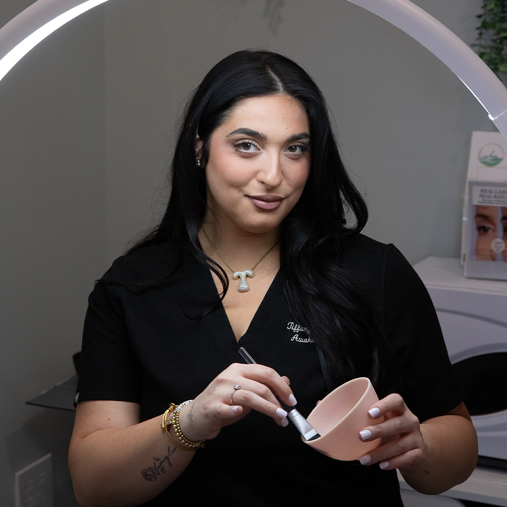 A woman esthetician in black scrubs mixing a facial mask in a pink bowl with a brush under a ring light in a clinic.